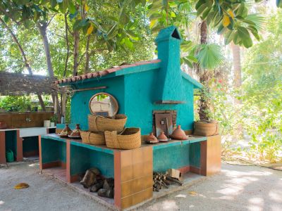 Zona de cocción exterior con horno turquesa y chimenea, encimeras de azulejos terracota y leña almacenada, enmarcada por exuberante vegetación tropical.