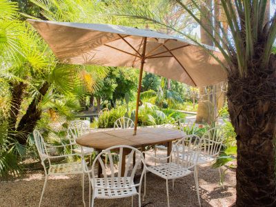 Zona de comedor exterior con mesa redonda de madera y seis sillas de metal blanco bajo una gran sombrilla beige. Rodeado de exuberantes palmeras tropicales y suelo de grava, con un estanque visible al fondo.
