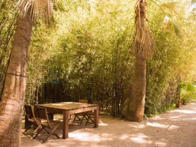 Zona de comedor exterior privada con una robusta mesa de madera y sillas plegables, ubicada en un sendero de gravilla entre palmeras y un denso seto de bambú con luces de guirnalda.