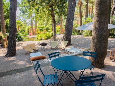 Zona de estar exterior con mesa bistro azul y sillas de tela sobre grava, sombreada por árboles con luces de guirnalda, junto a un estanque.