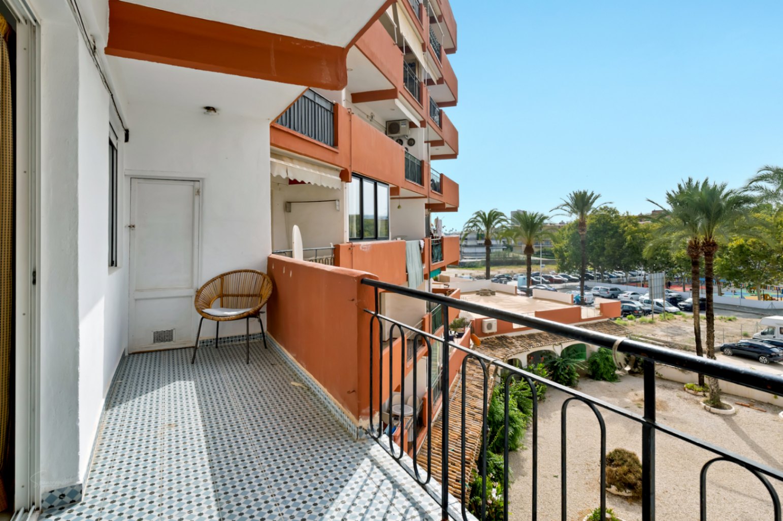 Balcón soleado con azulejos geométricos azules y blancos, equipado con una silla de mimbre y puerta de trastero. Vistas a palmeras y aparcamiento residencial.