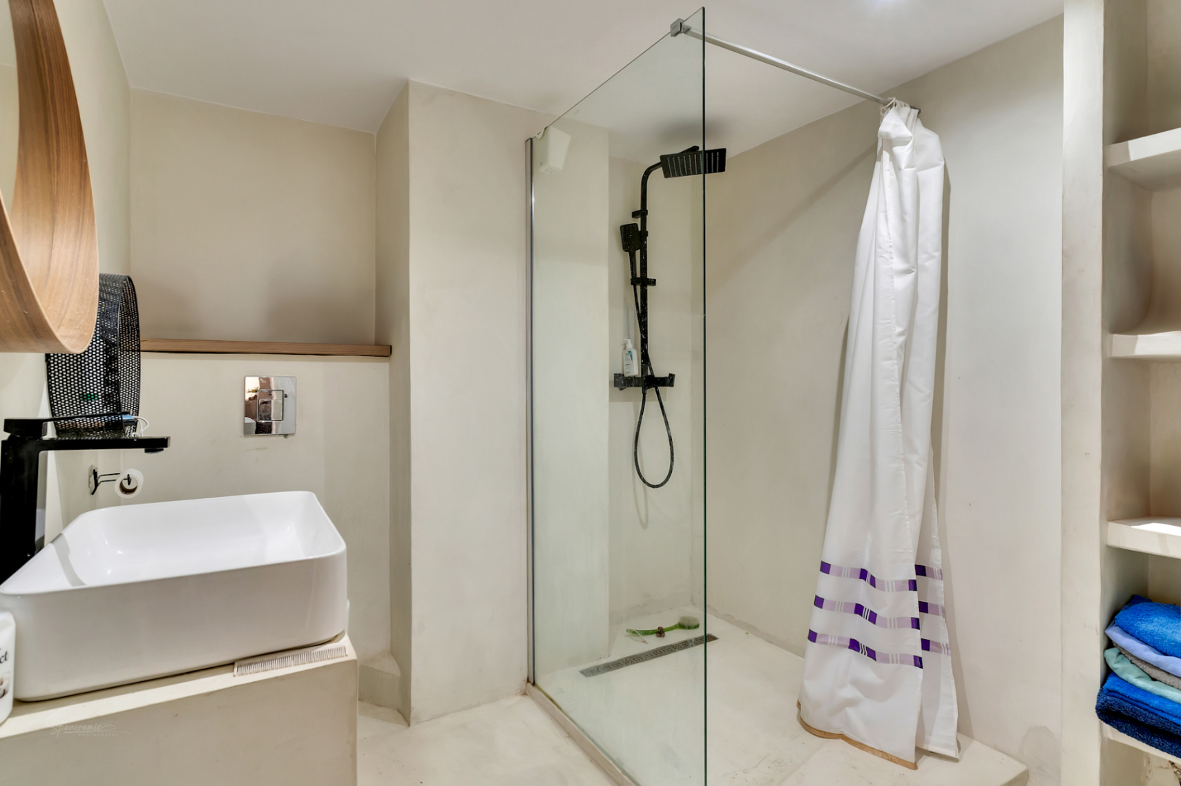 Baño moderno con lavabo sobre encimera, grifería negra mate y ducha a ras de suelo con mampara de cristal. Destacan el espejo de madera y estantes integrados.