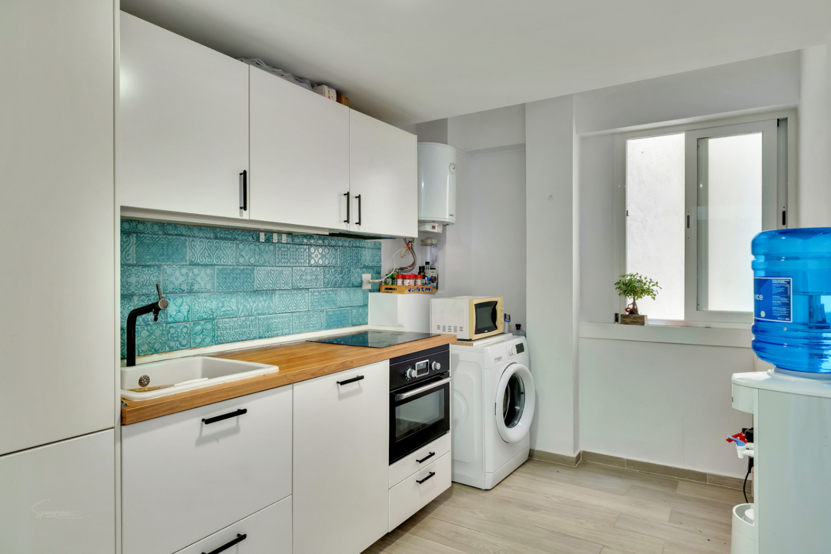 Cocina moderna con armarios blancos, encimera de madera clara y salpicadero de azulejos turquesa con patrón. Equipada con electrodomésticos integrados y zona de lavadero con lavadora.