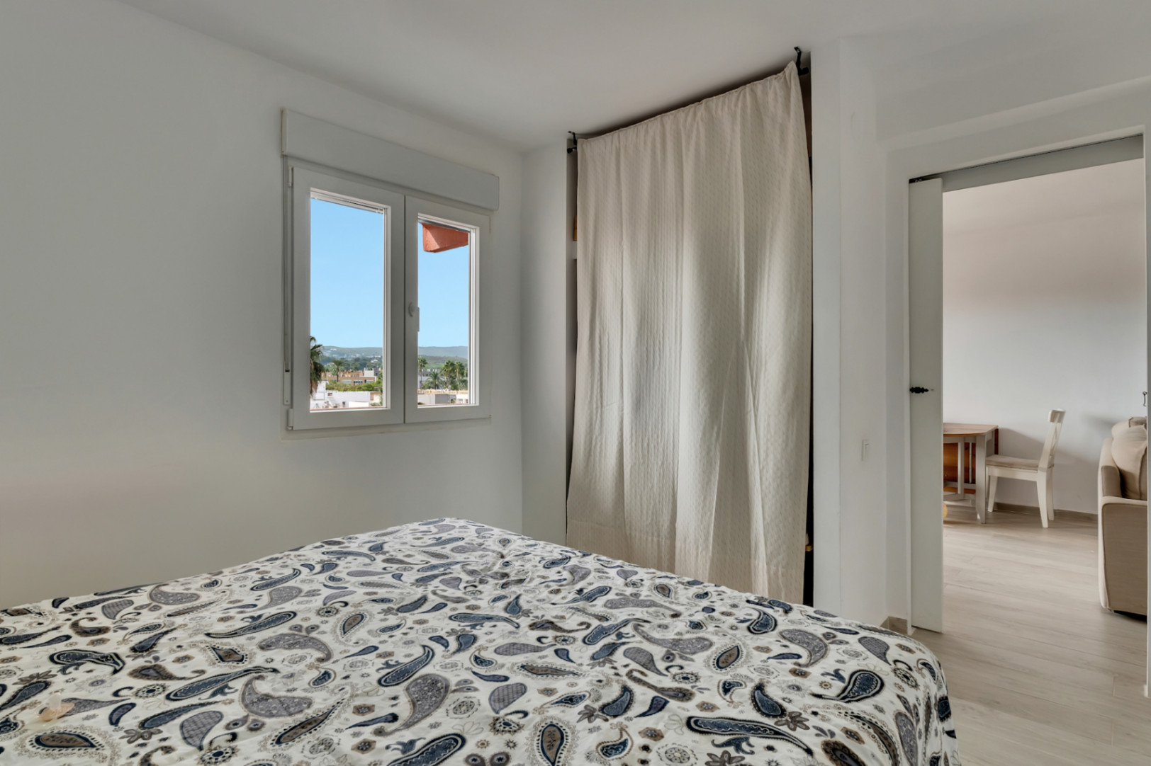 Dormitorio luminoso con cama de edredón estampado, ventana con vistas urbanas y un discreto armario cubierto por una cortina clara, con acceso a otra estancia.
