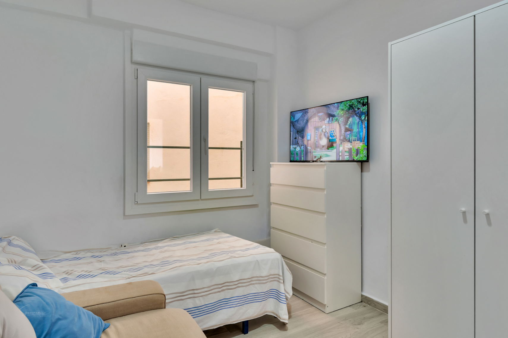 Dormitorio secundario con cama individual, armario empotrado blanco y cómoda con TV. La ventana de doble hoja proporciona luz natural.
