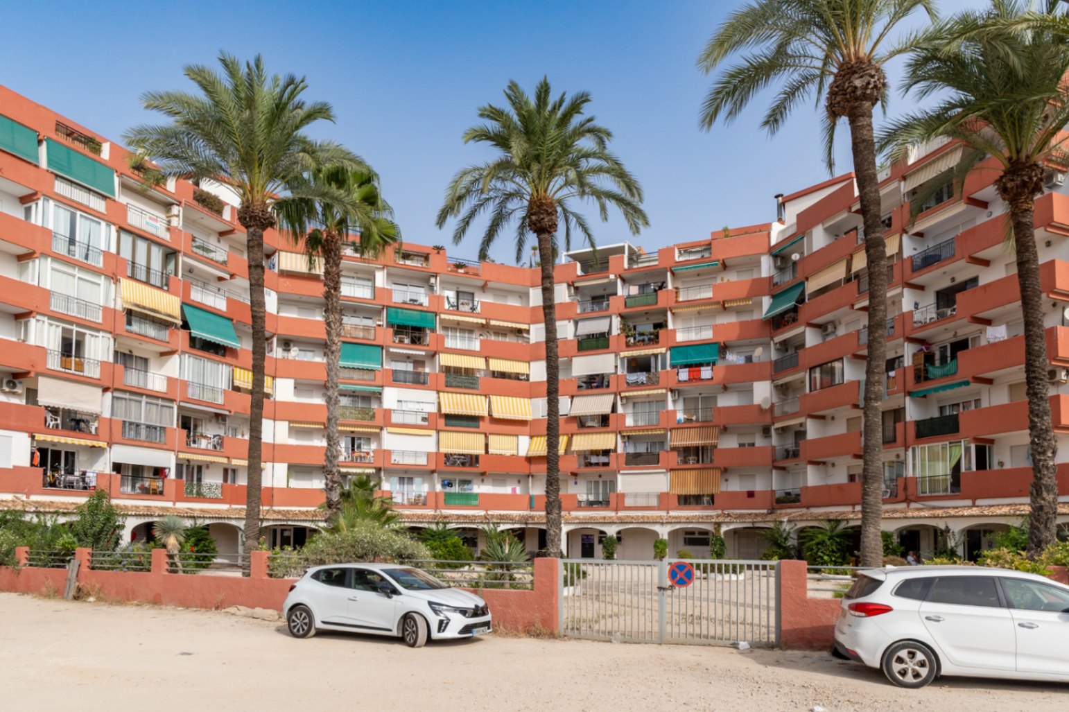 Edificio de apartamentos curvo con fachada terracota y balcones con toldos variados. Palmeras altas en primer plano y zona de aparcamiento exterior con puerta motorizada.