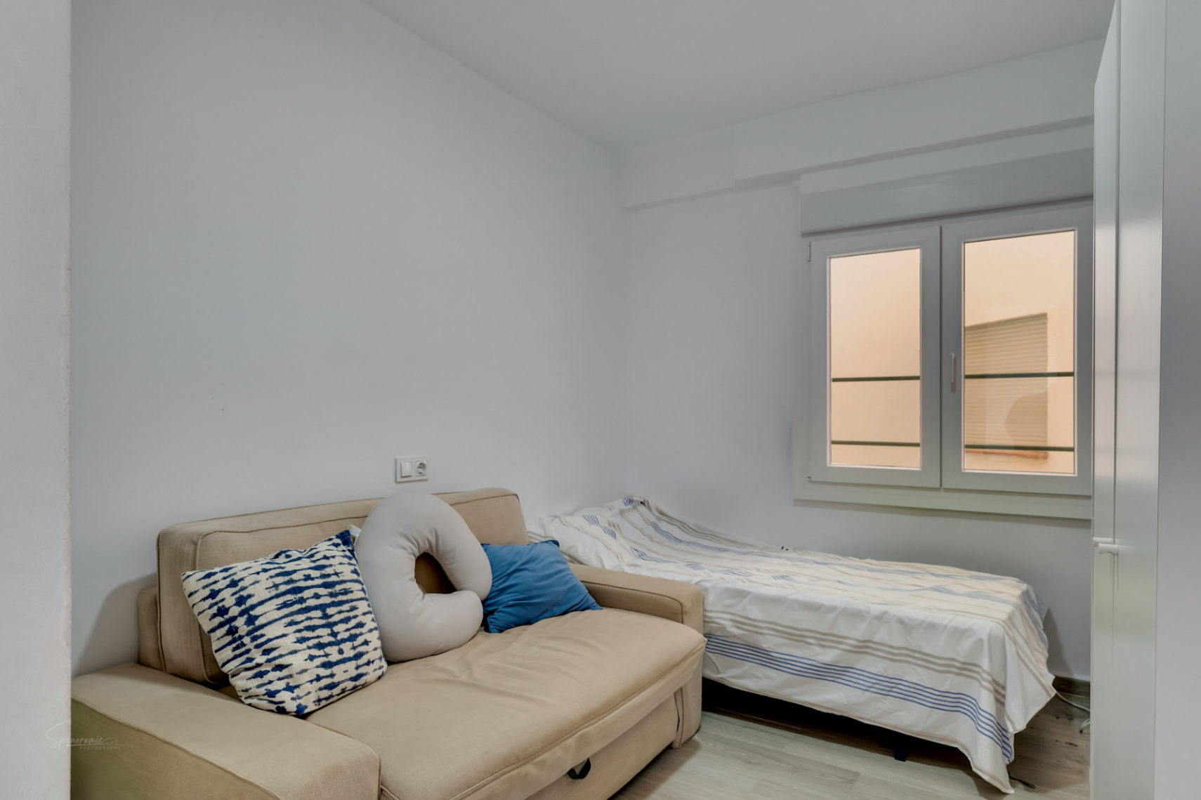 Habitación auxiliar con sofá cama beige y cama individual, paredes blancas y ventana que aporta luz natural difusa.
