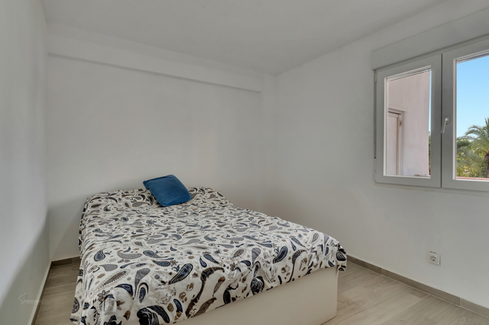 Luminoso dormitorio con paredes blancas y suelo laminado claro. Cama con edredón de estampado paisley y cojín azul, junto a una ventana con luz natural y vistas a palmeras.