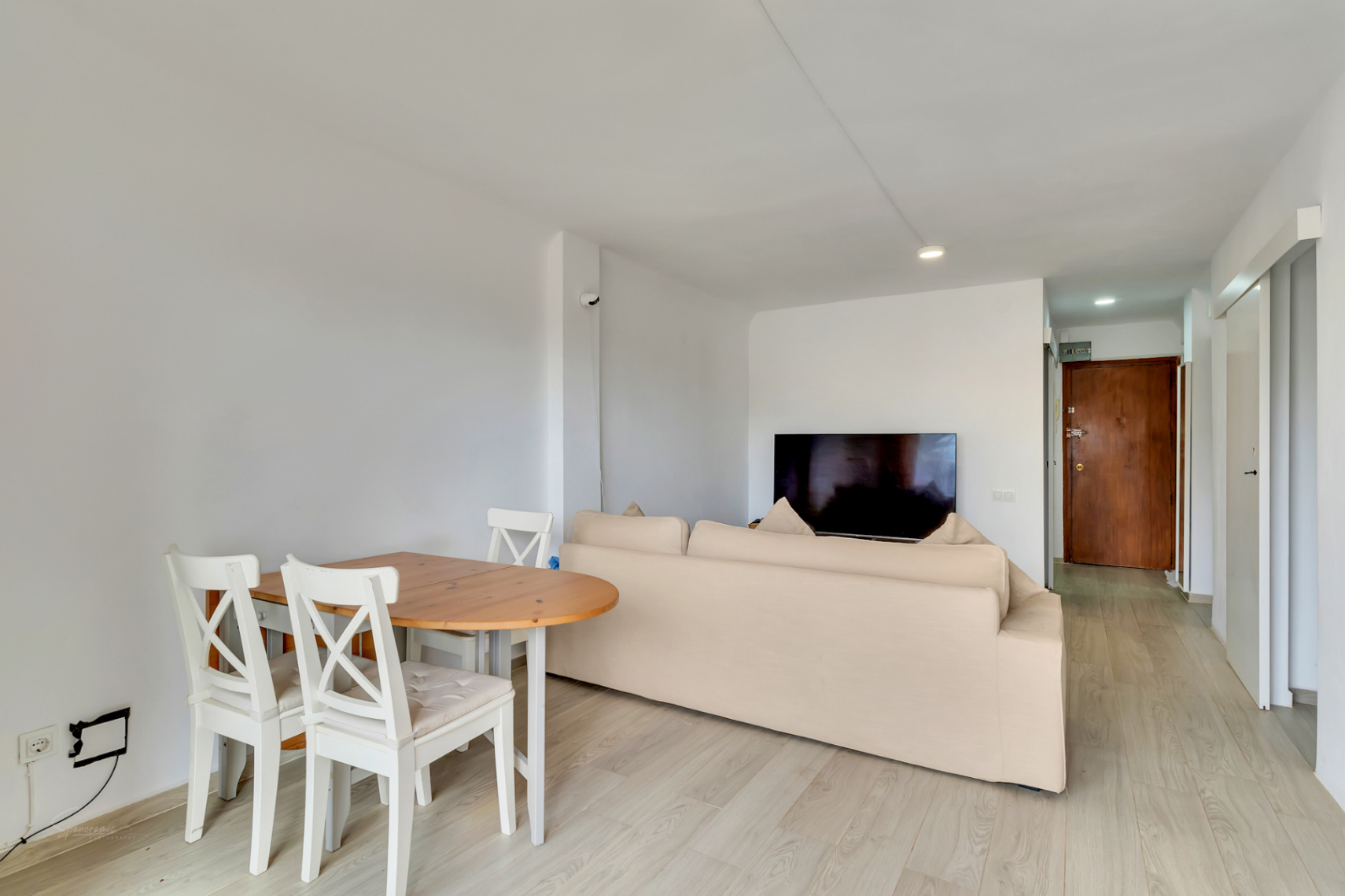 Salón-comedor diáfano con mesa de madera clara y sillas blancas, sofá beige y televisor. Suelo laminado claro y puerta de entrada de madera oscura al fondo.