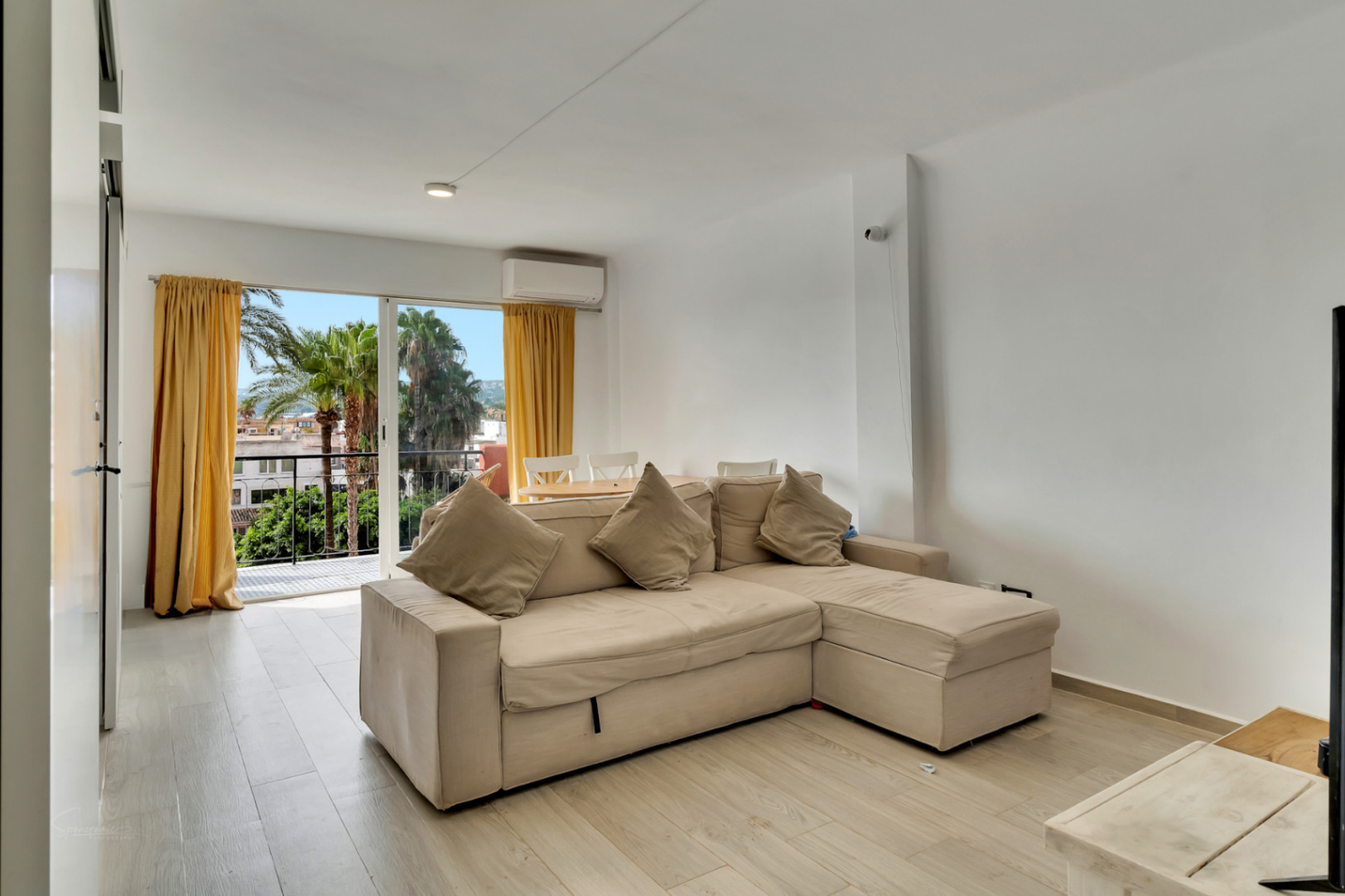 Salón luminoso con sofá beige en L, suelo de efecto madera clara y acceso a balcón con vistas a palmeras y cortinas amarillas.