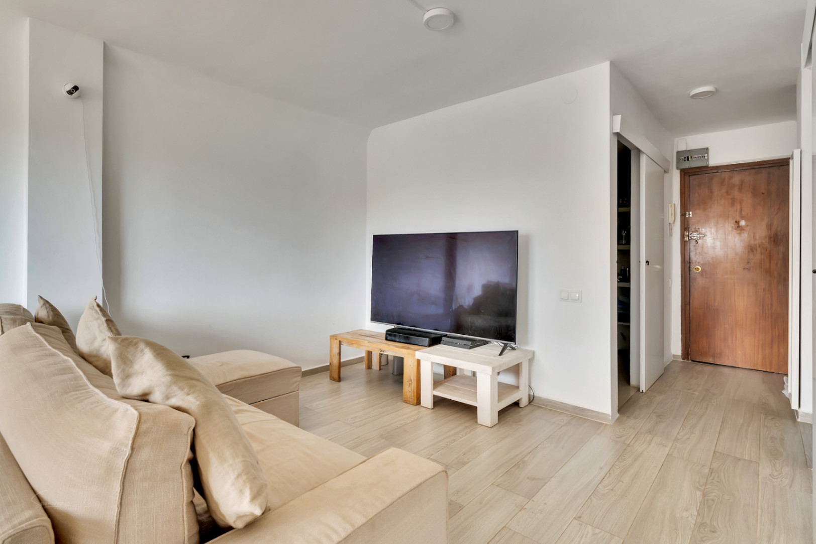 Salón luminoso con sofá seccional beige y TV sobre mesas de madera clara. El suelo laminado claro se extiende hacia la puerta de entrada de madera oscura y un espacio con estantes.