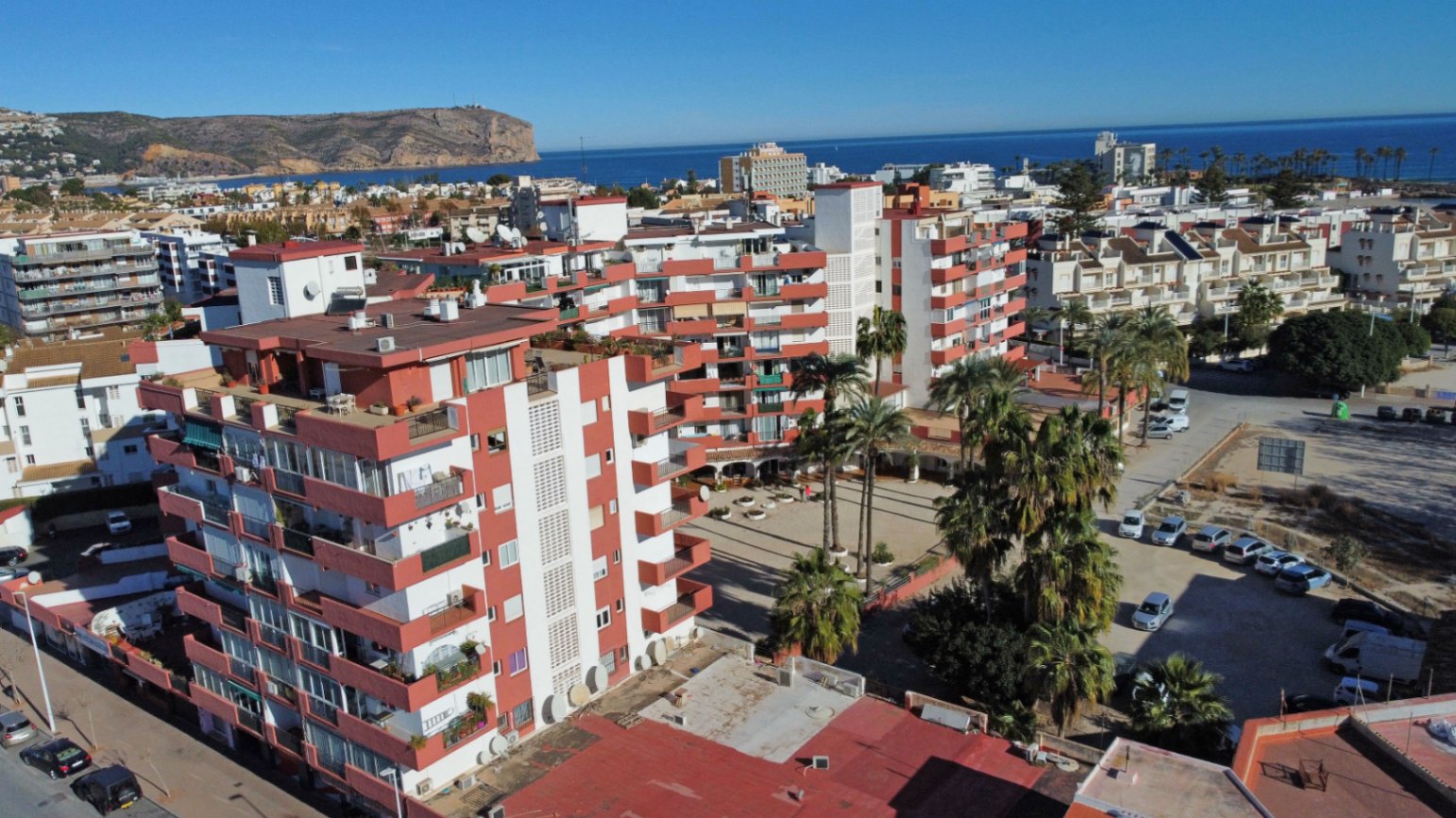 Vista aérea del complejo residencial con edificios de fachadas blancas y balcones rojizos, palmeras y el mar Mediterráneo al fondo.