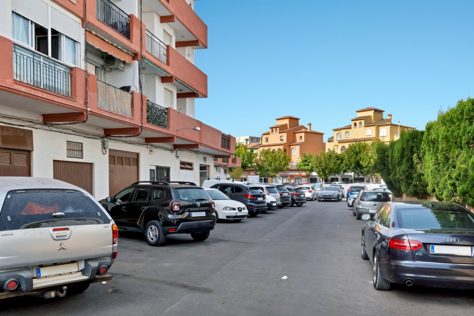 Zona de aparcamiento exterior con varios vehículos, flanqueada por un edificio residencial de balcones naranjas y estructuras tradicionales ocres al fondo bajo un cielo despejado.
