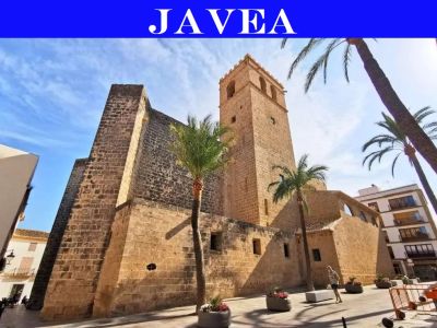 Antigua iglesia de piedra con torre campanario en Jávea, destacando su robusta mampostería y contrafuertes, en una plaza soleada con palmeras.