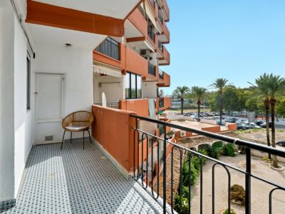 Balcón soleado con azulejos geométricos azules y blancos, equipado con una silla de mimbre y puerta de trastero. Vistas a palmeras y aparcamiento residencial.