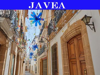 Callejón estrecho en el centro histórico de Jávea, con puertas de madera enmarcadas en piedra, balcones de hierro forjado y adornos festivos aéreos con formas de estrellas azules.
