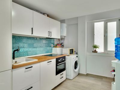 Cocina moderna con armarios blancos, encimera de madera clara y salpicadero de azulejos turquesa con patrón. Equipada con electrodomésticos integrados y zona de lavadero con lavadora.
