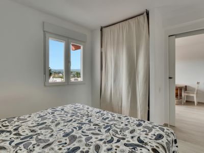 Dormitorio luminoso con cama de edredón estampado, ventana con vistas urbanas y un discreto armario cubierto por una cortina clara, con acceso a otra estancia.
