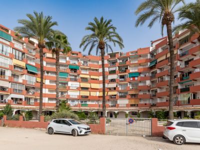 Edificio de apartamentos curvo con fachada terracota y balcones con toldos variados. Palmeras altas en primer plano y zona de aparcamiento exterior con puerta motorizada.