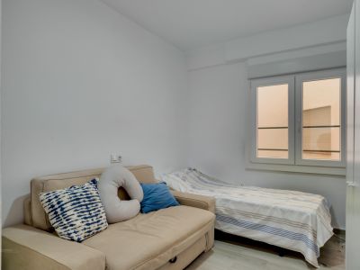 Habitación auxiliar con sofá cama beige y cama individual, paredes blancas y ventana que aporta luz natural difusa.