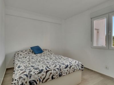 Luminoso dormitorio con paredes blancas y suelo laminado claro. Cama con edredón de estampado paisley y cojín azul, junto a una ventana con luz natural y vistas a palmeras.