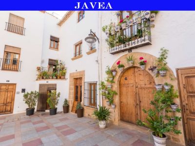 Rincón pintoresco con fachadas blancas, puertas de madera rústicas y balcones con barandillas de hierro forjado. Numerosas macetas con flores adornan las paredes y el suelo de baldosas rojas y grises.