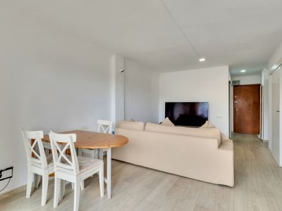 Salón-comedor diáfano con mesa de madera clara y sillas blancas, sofá beige y televisor. Suelo laminado claro y puerta de entrada de madera oscura al fondo.