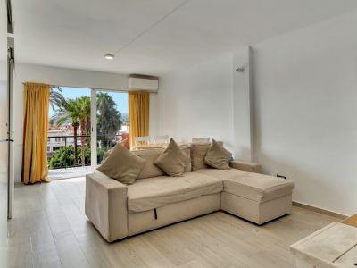 Salón luminoso con sofá beige en L, suelo de efecto madera clara y acceso a balcón con vistas a palmeras y cortinas amarillas.