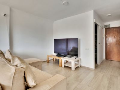Salón luminoso con sofá seccional beige y TV sobre mesas de madera clara. El suelo laminado claro se extiende hacia la puerta de entrada de madera oscura y un espacio con estantes.