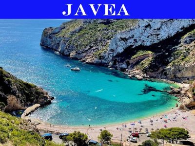 Vista aérea de una cala de Jávea con aguas turquesas y cristalinas, una playa de guijarros con bañistas y acantilados rocosos cubiertos de vegetación mediterránea.