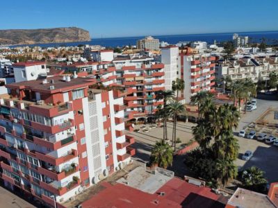 Vista aérea del complejo residencial con edificios de fachadas blancas y balcones rojizos, palmeras y el mar Mediterráneo al fondo.