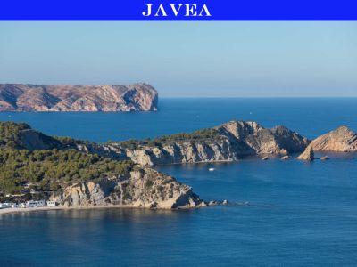 Vista panorámica de la costa escarpada de Jávea, con acantilados cubiertos de pinos, una cala con casas blancas y pequeños islotes rocosos en el Mediterráneo azul, y un faro distante en un cabo.
