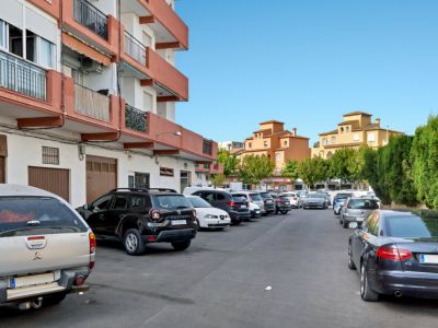 Zona de aparcamiento exterior con varios vehículos, flanqueada por un edificio residencial de balcones naranjas y estructuras tradicionales ocres al fondo bajo un cielo despejado.