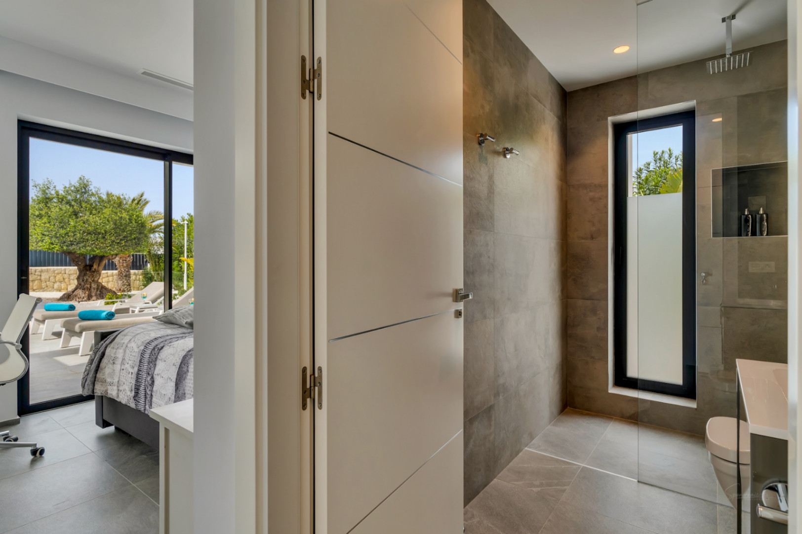 Acceso a un moderno baño con ducha de obra y ventana esmerilada, junto a un dormitorio con salida directa a terraza y vistas a un olivo. Suelos y paredes de gres en tonos neutros.