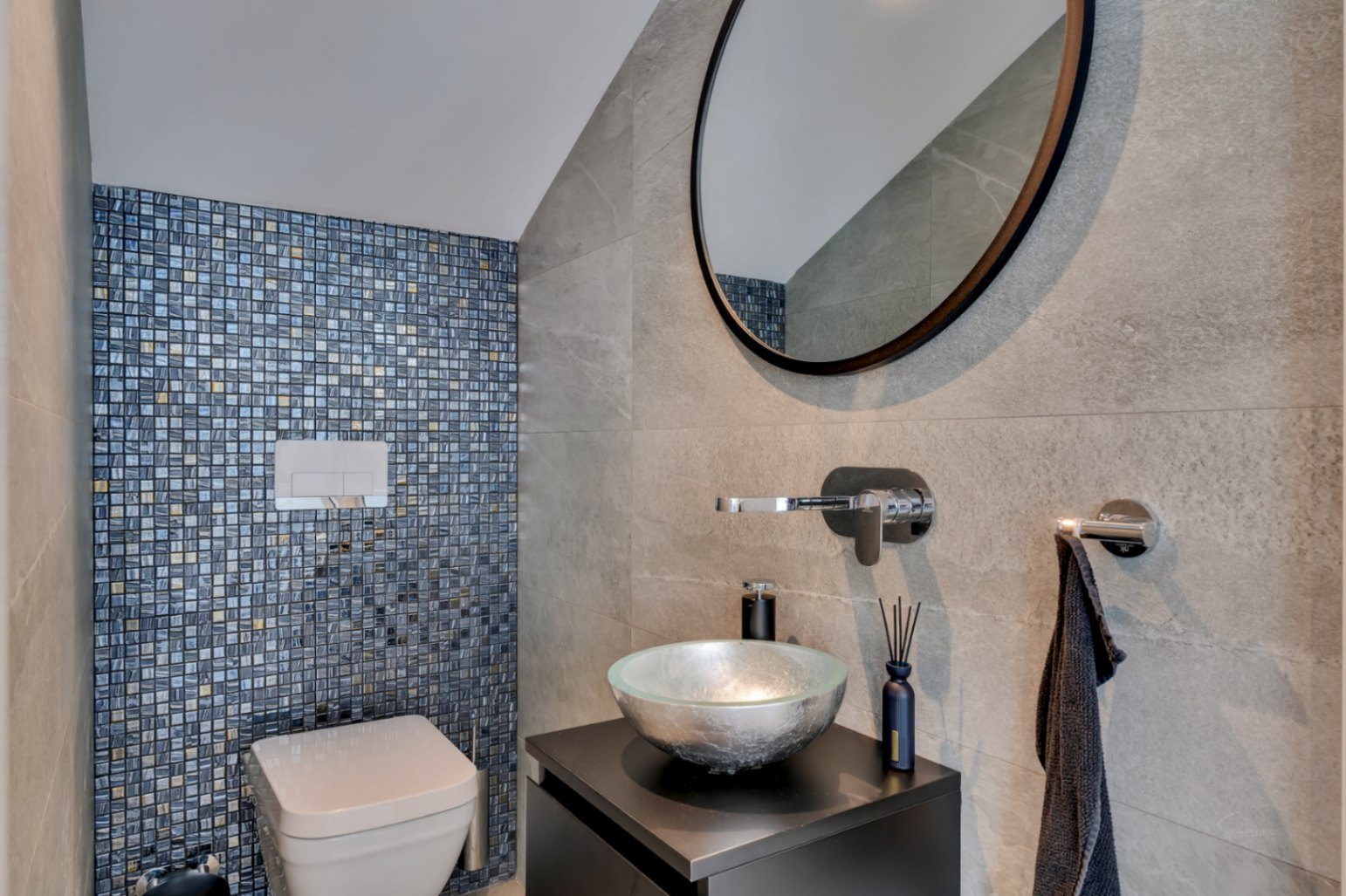 Aseo de cortesía con inodoro suspendido y pared de mosaico en tonos azules y dorados. Lavabo de cristal plateado sobre mueble oscuro y grifería mural.
