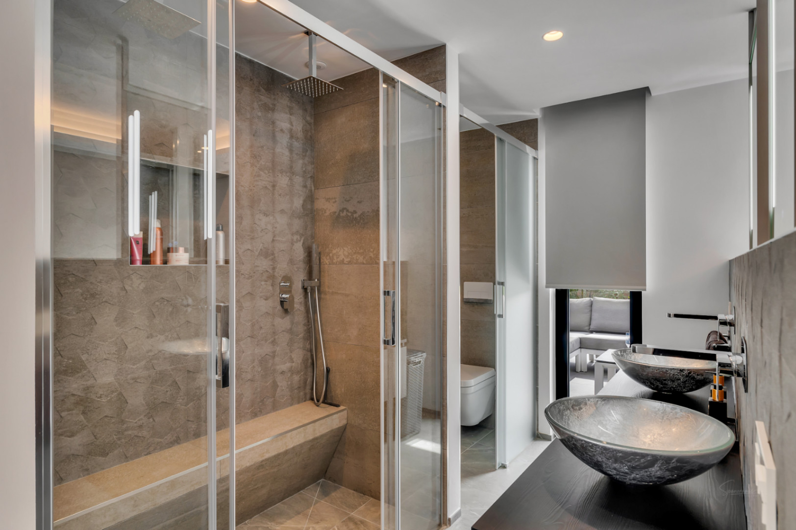 Baño moderno con ducha de obra acristalada, banco integrado y azulejos texturizados. Doble lavabo de cristal sobre encimera oscura y vista a terraza exterior.