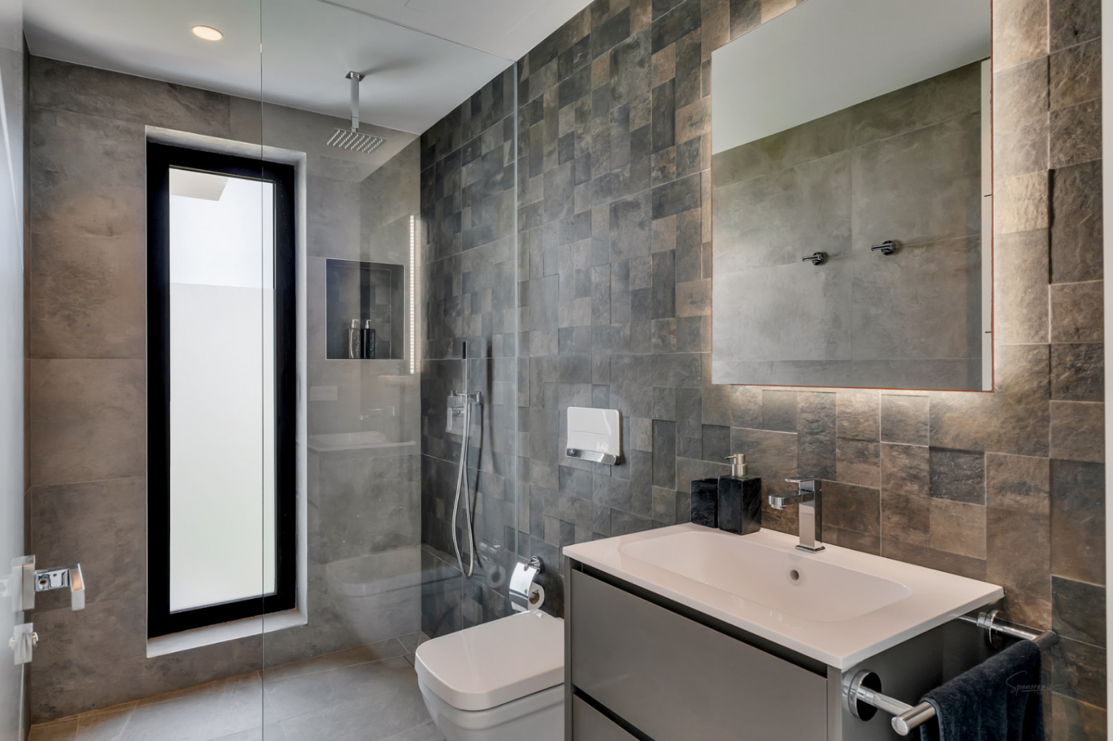 Baño moderno con ducha de obra, ventana vertical y azulejos grises texturizados. Lavabo flotante con espejo retroiluminado y sanitarios contemporáneos.