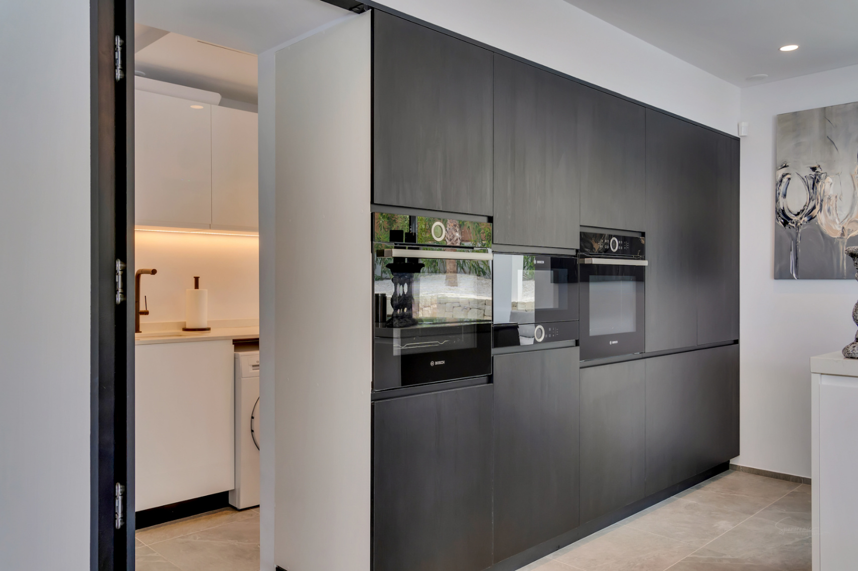 Cocina de concepto abierto con elegantes armarios oscuros integrados y electrodomésticos Bosch, conectada discretamente a una despensa funcional con lavadora y grifo cobrizo.