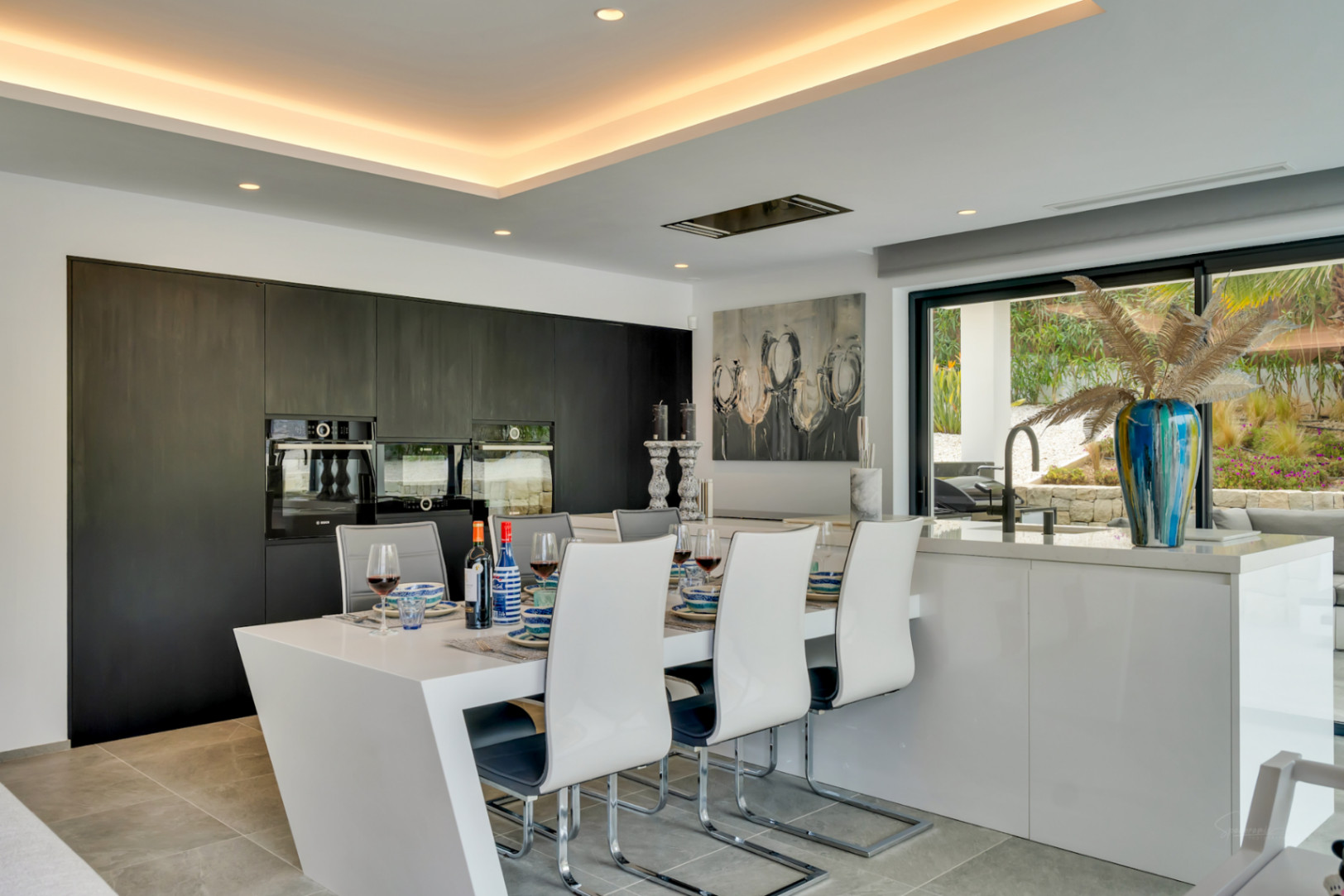 Cocina moderna de concepto abierto con isla-comedor integrada, armarios oscuros y hornos empotrados, conectada a un jardín a través de grandes ventanales.