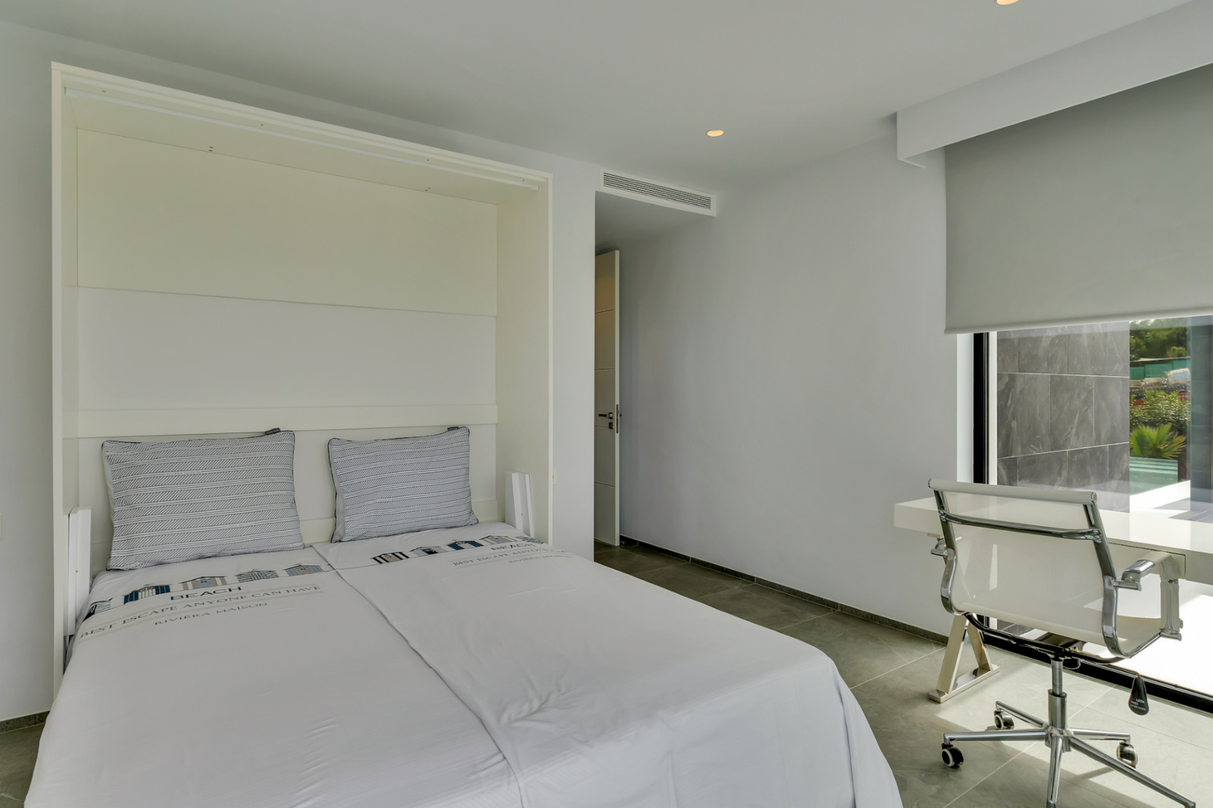 Dormitorio moderno con cama abatible integrada, escritorio minimalista junto a la ventana con vistas a la piscina y suelo de gres gris.