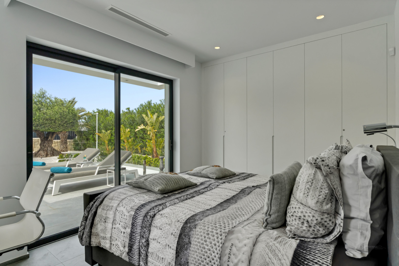 Dormitorio moderno con grandes puertas correderas de cristal que acceden a una terraza privada con tumbonas y exuberante vegetación mediterránea. Armarios empotrados integrados.