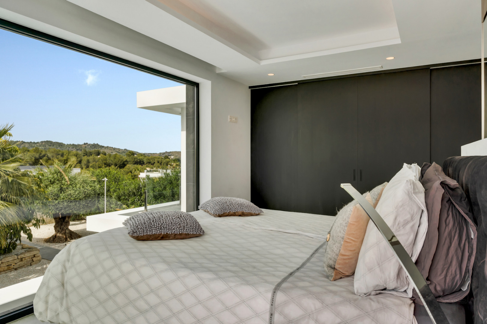 Dormitorio principal con ventanal de suelo a techo, ofreciendo vistas panorámicas a la vegetación y las colinas. Armarios empotrados oscuros y cama moderna con ropa de cama clara.