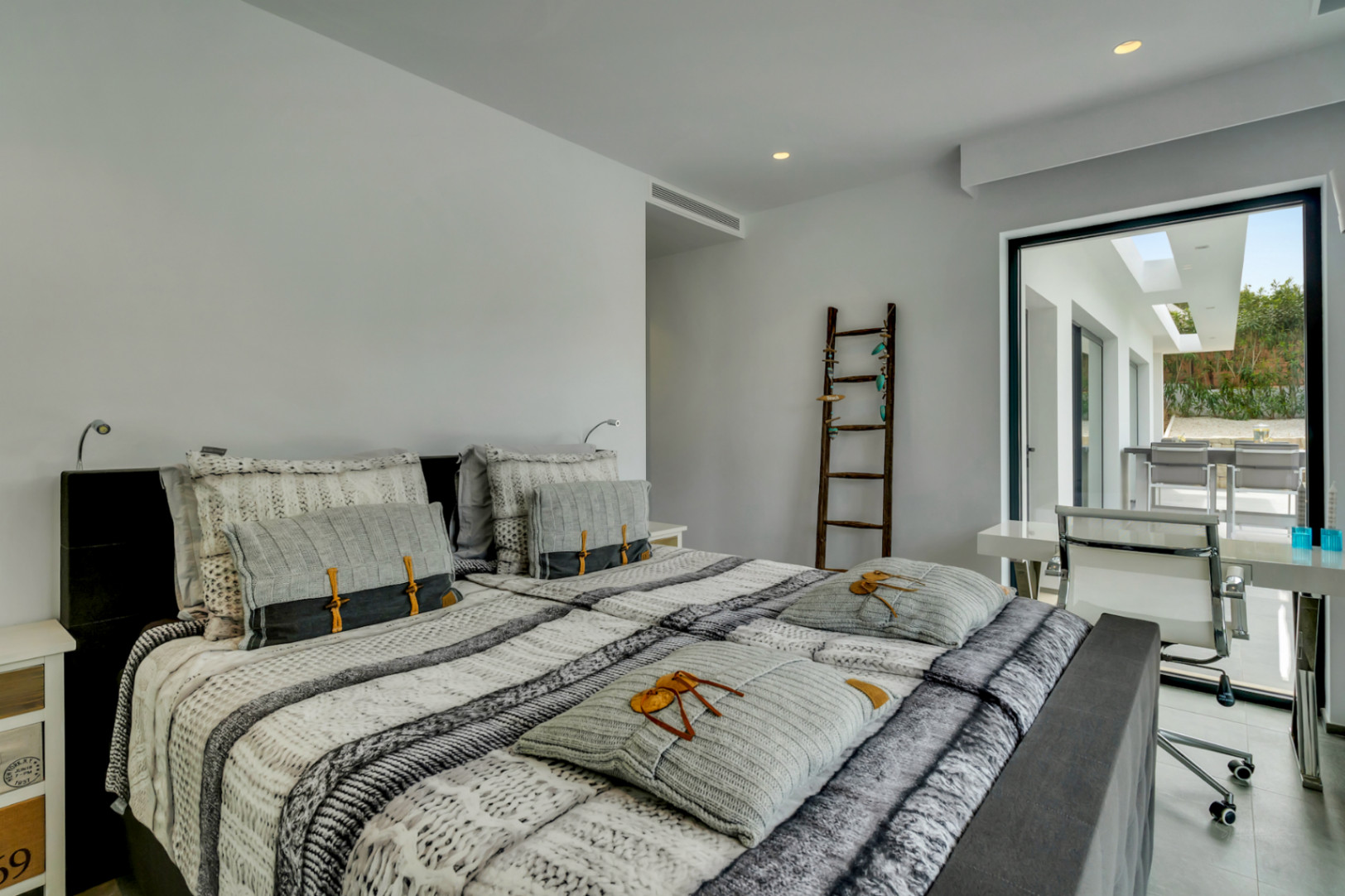 Dormitorio principal moderno con cama doble, acceso directo a una terraza exterior y área de trabajo integrada. Destacan la iluminación empotrada y una escalera decorativa.