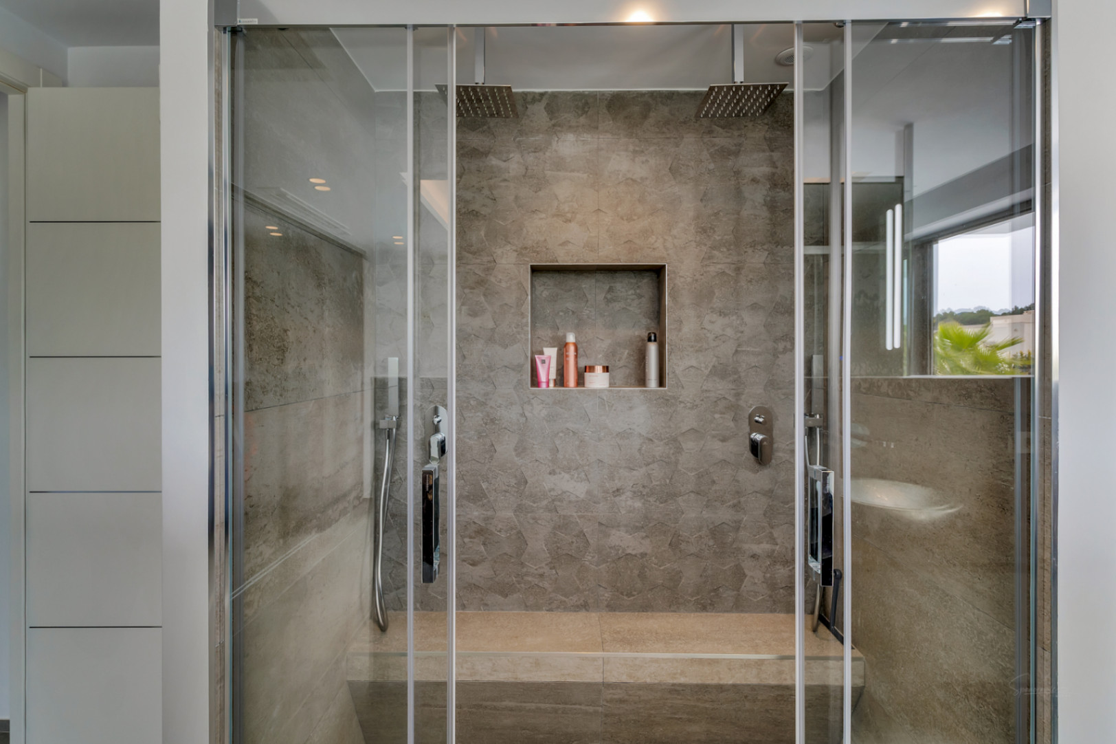Ducha moderna con mampara de cristal, azulejos de efecto piedra gris y dos rociadores de techo. Incluye banco integrado, nicho para productos y armarios blancos. Vista exterior con vegetación.