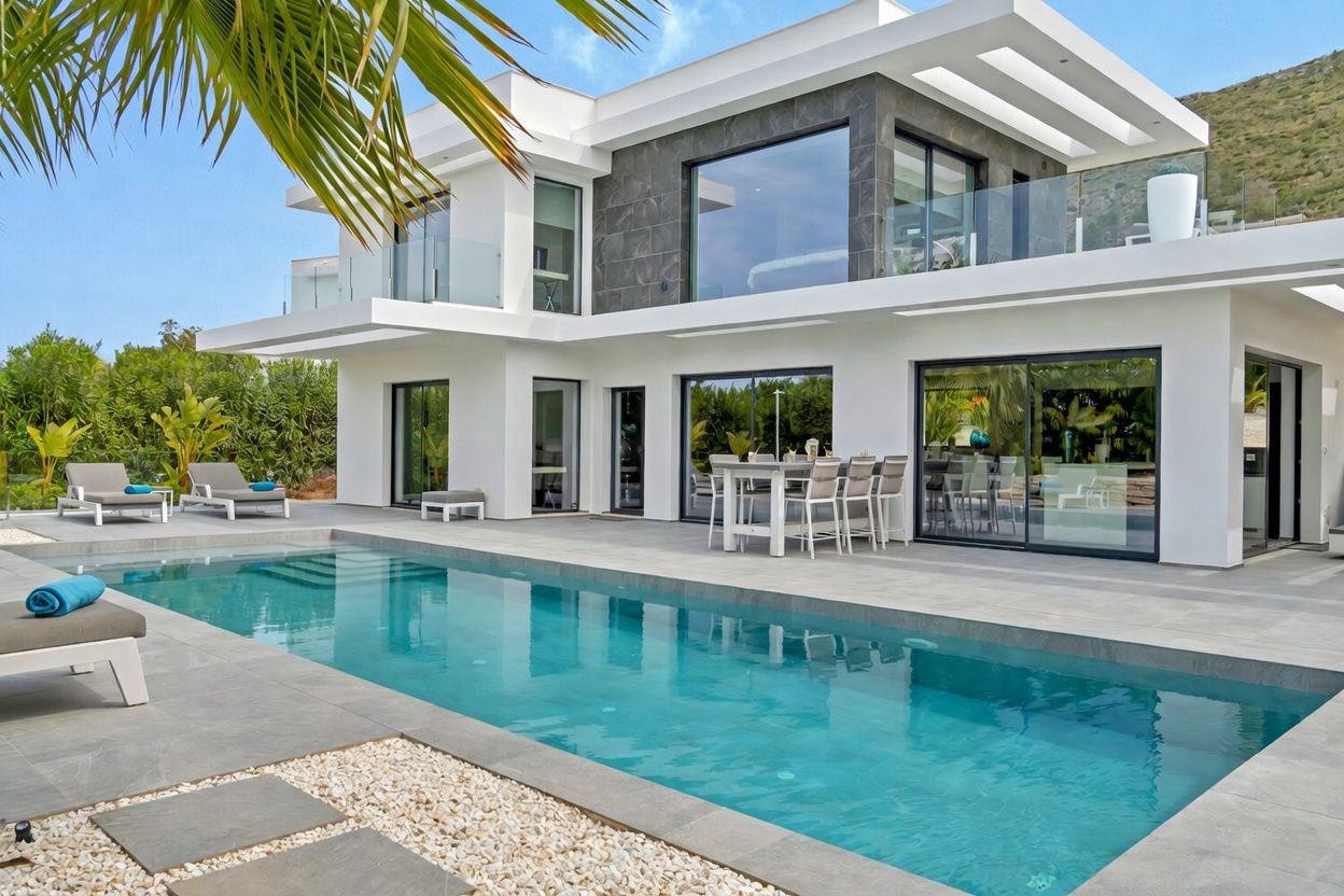 Exterior de villa moderna blanca con piscina rectangular y patio de baldosas grises. Comedor exterior junto a puertas de cristal, balcón acristalado y detalles de fachada en gris oscuro en el nivel superior.