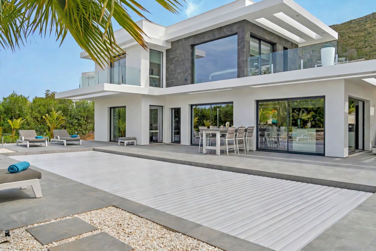 Exterior de villa moderna con piscina cubierta por lamas blancas y amplia terraza de gres gris. Incluye zona de comedor exterior y grandes ventanales que reflejan el entorno.