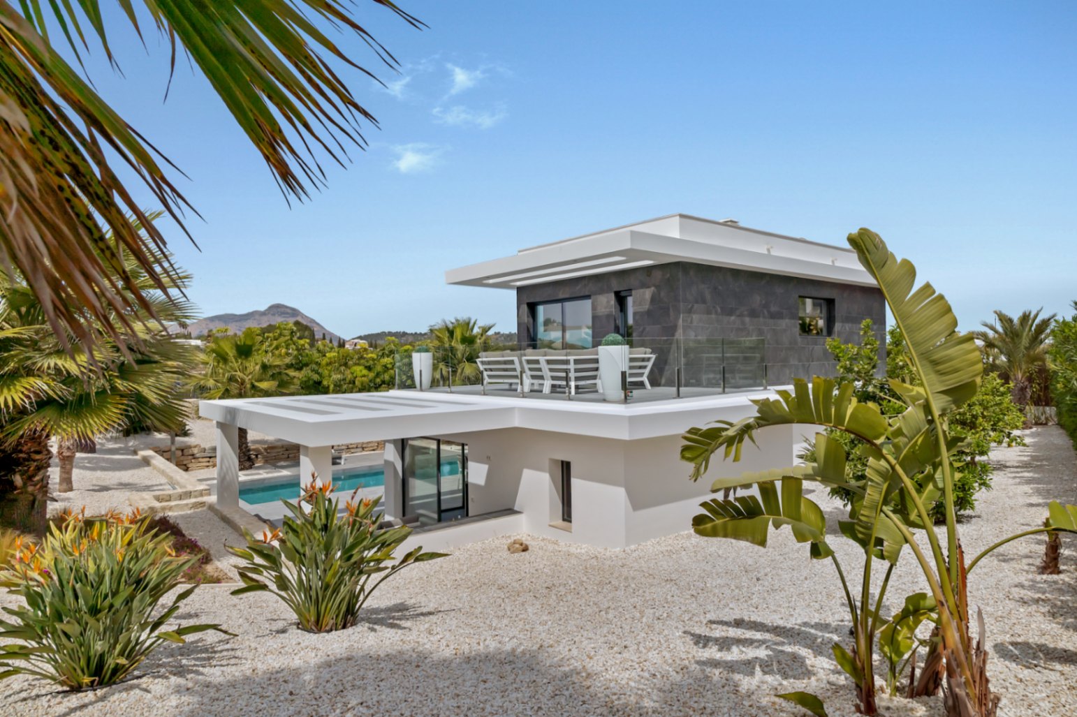 Exterior de villa moderna de varios niveles con revestimiento blanco y piedra oscura. Destaca una terraza superior con barandillas de cristal y asientos, rodeada de grava blanca y plantas tropicales.