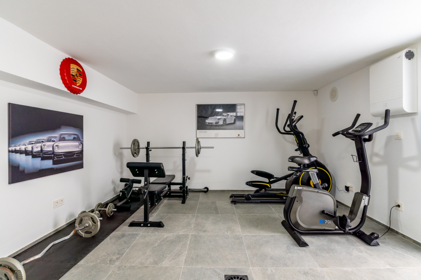 Gimnasio doméstico con banco de pesas, elíptica y bicicleta estática, sobre suelo de baldosas grises y decorado con arte automovilístico de Porsche.