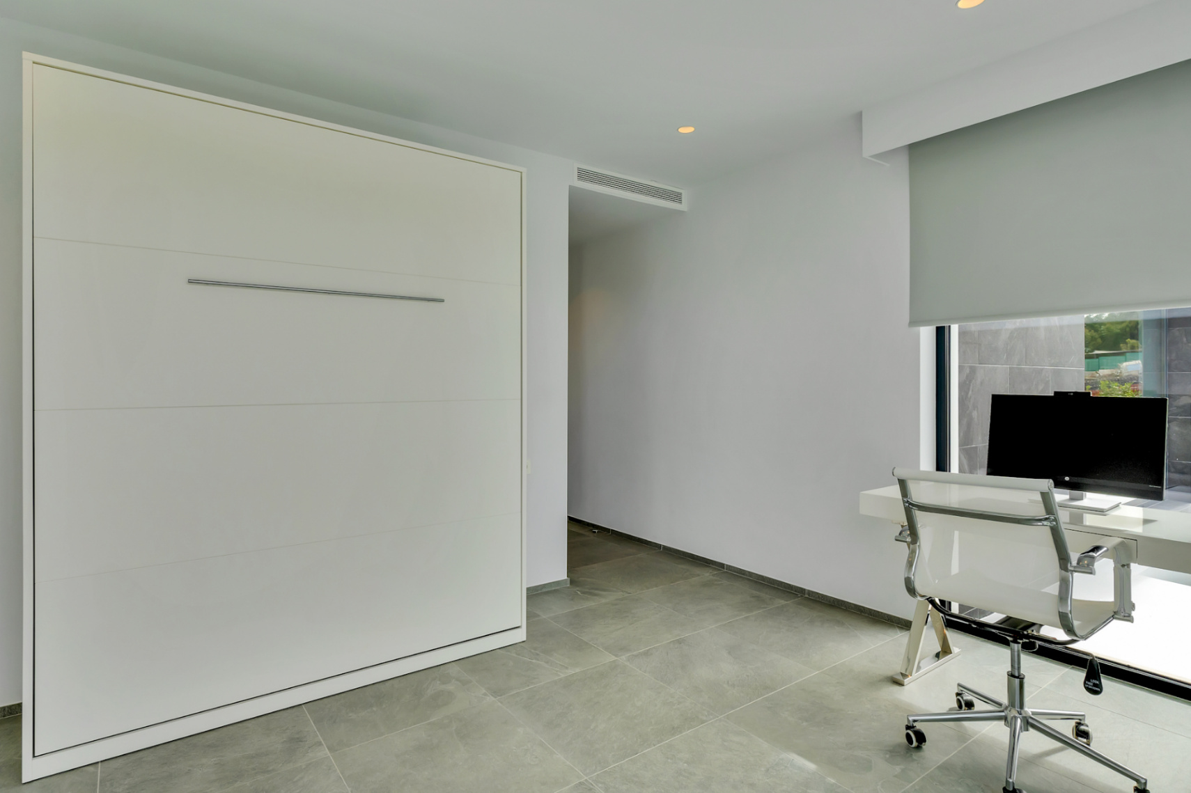 Habitación versátil con cama abatible integrada y escritorio minimalista. Suelo de gres gris claro, ventana con persiana enrollable y vistas al exterior.