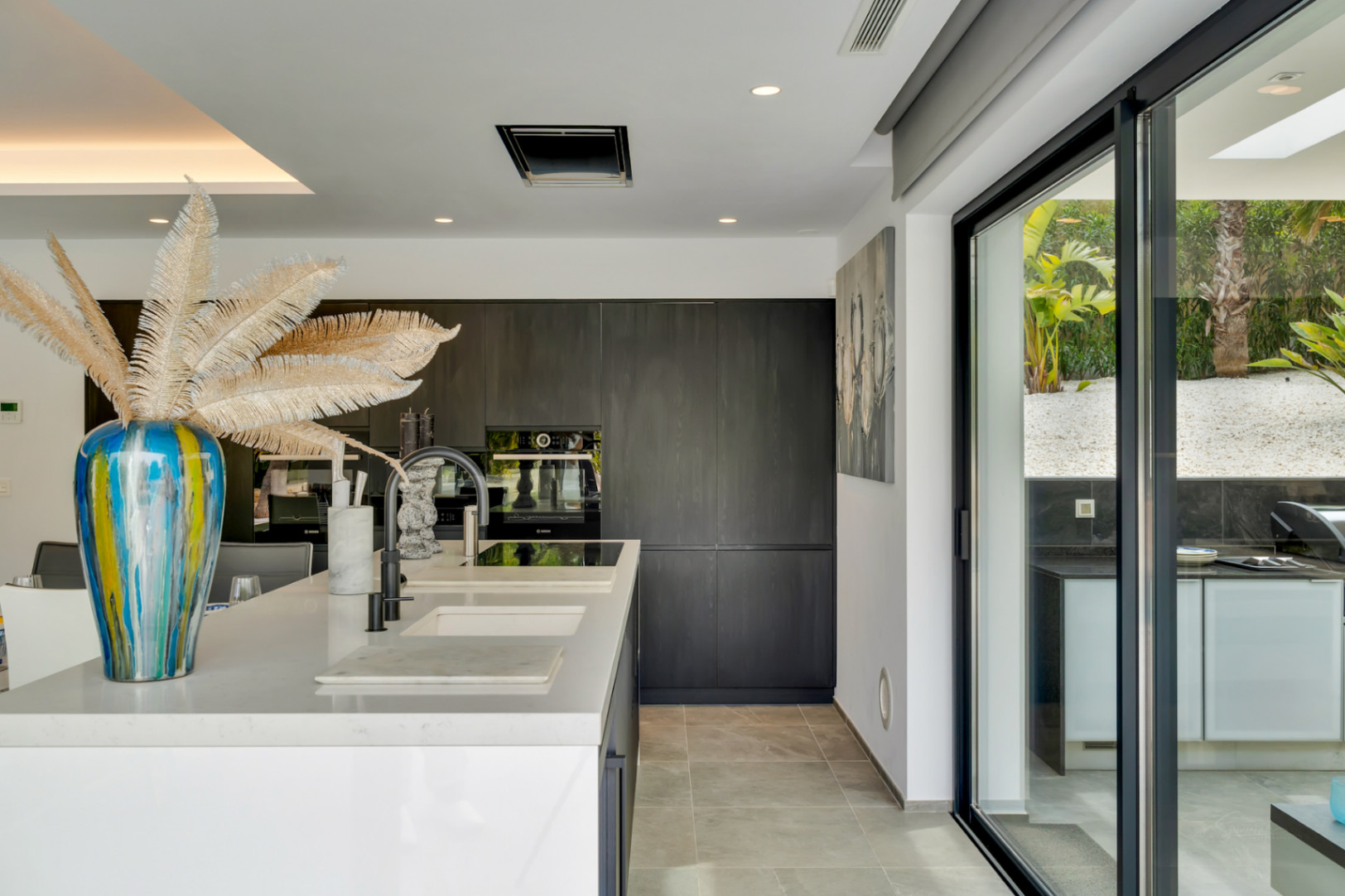 Isla de cocina moderna con encimera blanca, fregaderos integrados y grifo negro. Armarios oscuros minimalistas con electrodomésticos empotrados, y puertas correderas de cristal hacia la cocina exterior y jardín.